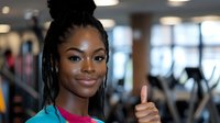 Young African American woman with dreadlocks hairstyle showing thumbs up at fitness center, wearing turquoise sportswear, expressing positivity and confidence.