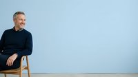 Middle-aged Caucasian man with gray hair wearing dark navy sweater, sitting on wooden stool against light blue background. Portrait for business, corporate, lifestyle, and professional use.