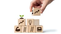 Hand placing wooden cube with growth arrow on stack of business concept blocks including lightbulb, ladder, and briefcase symbols on white background.