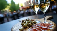 Two glasses of white wine served with Mediterranean appetizer platter featuring prosciutto, green olives on wooden board against blurred outdoor restaurant background.
