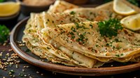 Homemade thin crepes or pancakes with herbs and spices served on rustic plate with lemon slices, fresh parsley garnish on wooden table.