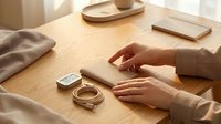 Woman's hands placing smartphone on wooden desk beside charging cable and earbuds case in cozy minimalist home workspace setting.