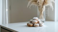Freshly baked sugar-dusted donuts stacked on white marble table with dried pampas grass in glass vase creating cozy minimalist kitchen scene.