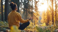 Young African American woman in yellow sweater meditating in lotus position during sunset in forest, peaceful mindfulness practice surrounded by nature.