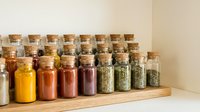Organized spice jars with cork lids arranged on wooden shelf, displaying colorful dried herbs, seasonings and spices for cooking, kitchen storage and pantry organization.