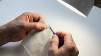 Senior craftsman hands carefully threading needle with white thread under bright lamp light for detailed sewing work and textile repair projects.