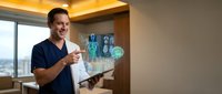 Smiling male doctor in blue scrubs using futuristic holographic medical interface technology in modern hospital room with digital patient data and diagnostic tools.