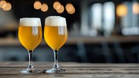 Two full glasses of unfiltered craft beer with foam head on wooden table against blurred bar background with bokeh lights, selective focus on cold beverages.