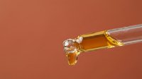 Golden oil or serum dripping from dropper bottle against warm terracotta background, suitable for cosmetics, skincare and wellness product marketing.