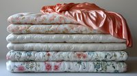 Neatly folded stack of vintage floral bedding linens and coral satin sheets arranged in soft natural lighting, showcasing delicate rose patterns and textures.