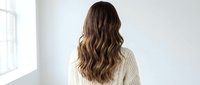 Young woman with long wavy brown hair wearing cream knit sweater standing by window in bright minimalist interior for lifestyle and fashion content.