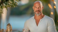 Confident mature Caucasian man with bald head and gray beard wearing white henley shirt smiling outdoors against blurred bokeh background with decorative lights.