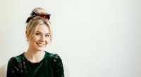 Young Caucasian woman with blonde hair wearing dark green velvet top and pink-tinted sunglasses pushed up on head, smiling against white background for portrait and lifestyle use.