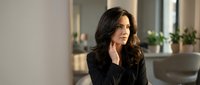 Professional businesswoman with dark wavy hair in black blazer sitting thoughtfully in modern office interior with natural lighting and plants.