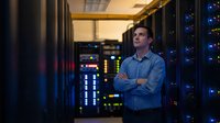 Confident Caucasian male IT administrator standing with crossed arms in modern server room with glowing blue lights and network equipment racks.