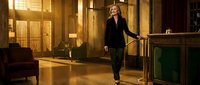 Professional blonde businesswoman in black suit walking confidently through modern office lobby with warm lighting and contemporary furniture.