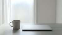 Minimalist workspace with laptop and coffee mug on white desk near bright window. Clean modern office setup for productivity and remote work concepts.