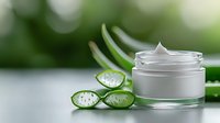 Fresh aloe vera slices with water drops next to white cosmetic cream jar on blurred green background, natural skincare and beauty concept.