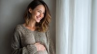 Young Caucasian pregnant woman with ombre hair smiling by window in cozy sweater, holding her baby bump with gentle expression of anticipation.