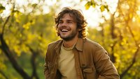 Young Caucasian man with beard and curly hair laughing outdoors in autumn park at golden hour sunset, wearing brown jacket and yellow t-shirt, expressing joy and happiness.