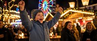 Young boy celebrating with raised arms at festive Christmas market with colorful ferris wheel and warm lights creating magical holiday atmosphere.