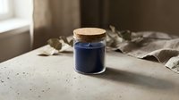 Dark blue candle in glass jar with cork lid on concrete surface near window with soft natural lighting for home decor and relaxation atmosphere.