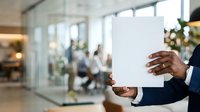 Businessman holding blank white document or certificate in modern office environment with colleagues in background, corporate workplace setting.