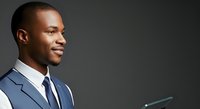 Young African American male professional in business suit and tie, holding tablet device, side profile portrait against neutral gray background for corporate and technology concepts.