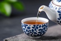 Pouring hot tea from traditional blue and white porcelain teapot into matching ceramic cup with floral pattern on dark background, close up view of steaming drink.