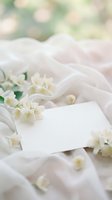 Blank white card surrounded by delicate white flowers and soft fabric with blurred green foliage background, suitable for wedding invitations, greeting cards, and event announcements.