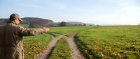 Farmer in brown jacket pointing at dirt road through green crop field on rural countryside landscape.