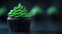 Dark chocolate cupcake with bright green buttercream frosting swirled on top, photographed against black background with soft bokeh effect and selective focus.