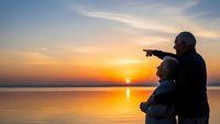 Father and son silhouettes pointing at sunset over calm lake water. Family bonding moment during golden hour with peaceful reflection on surface.