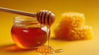 Golden honey dripping from wooden dipper onto spoon with glass jar and honeycomb pieces on vibrant yellow background. Perfect for food packaging or wellness blog.