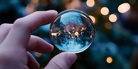 Magical crystal ball held in fingers reflects inverted winter landscape with bokeh lights, creating dreamy atmospheric scene against blurred evening background.