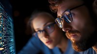 Two focused software developers analyzing code on computer screen in dark office environment with programming interface and digital data visualization.