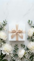 Elegant white gift box with champagne satin ribbon surrounded by peonies, eucalyptus, and baby's breath on marble surface.