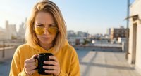 Young blonde woman in yellow hoodie and sunglasses holding coffee cup on urban rooftop terrace with city skyline background during golden hour sunrise.