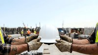 Construction worker holding white safety helmet on active building site with machinery and cityscape background for occupational safety and industrial workplace concepts.