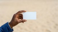 Person with dark skin tone holding blank white card at beach, wearing blue sleeve. Suitable for business cards, marketing mockups, travel promotions, and branding presentations.