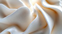 Soft cream-colored fabric with gentle folds and waves creating an elegant abstract background texture with subtle shadows and highlights in warm tones.