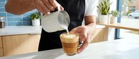 Professional barista pouring steamed milk into coffee cup creating latte art in modern cafe kitchen with natural lighting and plants in background.