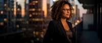 Young woman with curly dark hair and glasses wearing black blazer against blurred city skyline at sunset, suitable for corporate, business, and professional lifestyle content.
