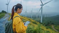 Young Asian female engineer in yellow jacket checking tablet at wind turbine farm on green hillside during cloudy day, sustainable energy concept.