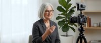 Mature woman with white hair and glasses wearing dark blazer gestures while recording video content on professional camera mounted on tripod in bright studio with potted plant and shelving.
