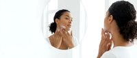 Young woman with dark skin and black hair examining facial skin in round mirror, applying skincare product or moisturizer for beauty routine and wellness.