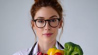 Young female nutritionist in white coat and glasses holding fresh vegetables and fruits against light background, promoting healthy eating and diet planning.