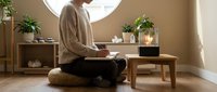 Young Asian woman meditating in peaceful home interior with plants and natural light streaming through round window creating zen atmosphere.