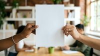 Two people holding blank white paper sheet in front of their faces indoors, suitable for advertising, marketing campaigns, presentations, and promotional materials.