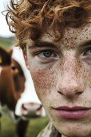 Close up portrait of young male with intense blue eyes, ginger curly hair and freckles on face, with cow in background on farm. Natural rural lifestyle.